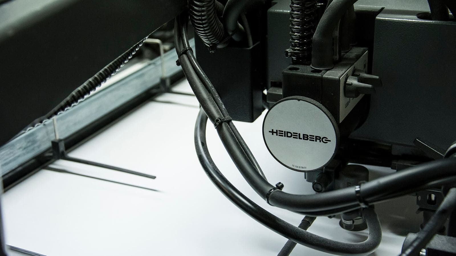 A modern offset press in close-up: the Heidelberg nameplate on a silver housing, with black hoses and cables above a sheet of white paper.