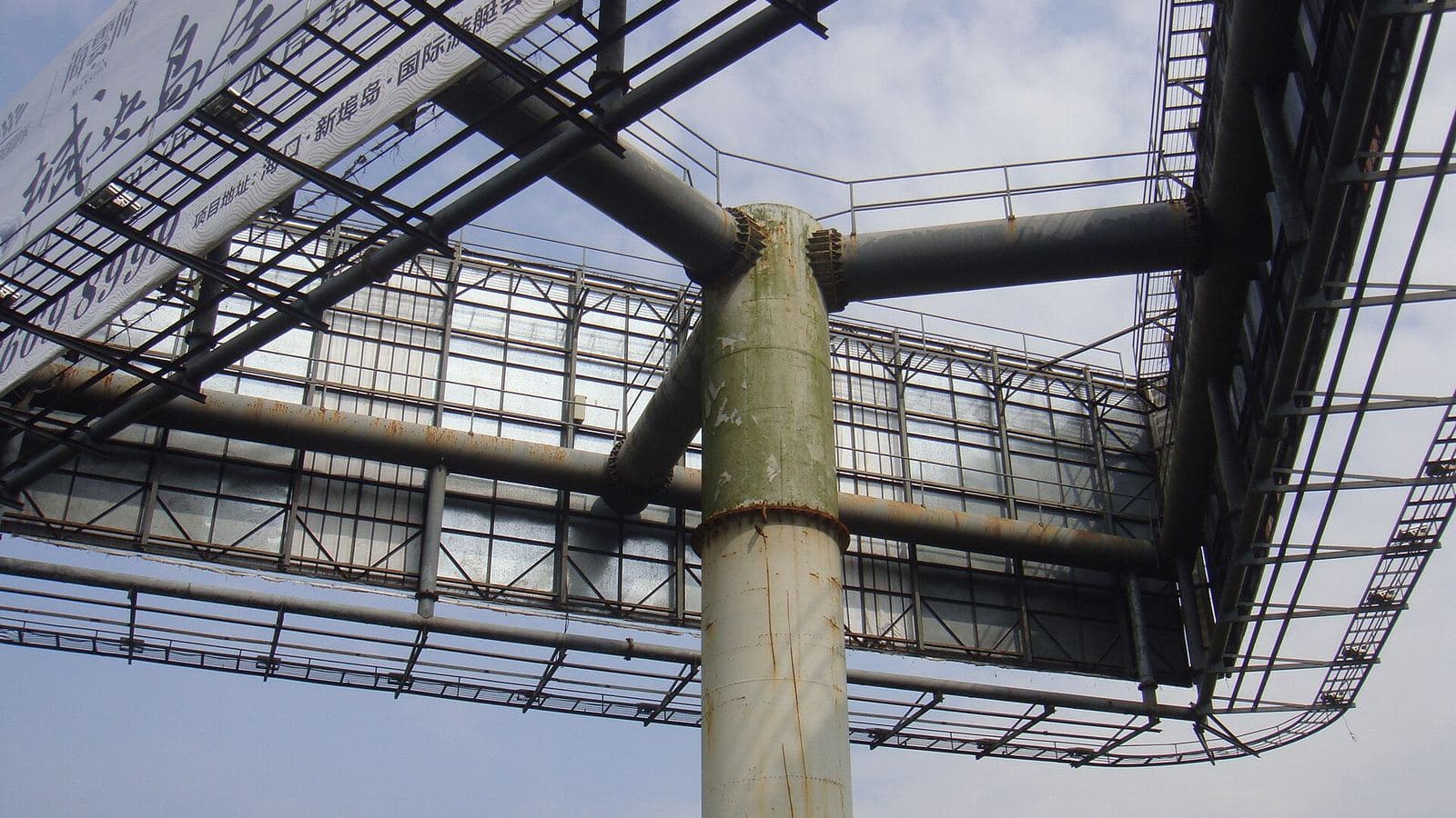 A steel billboard structure seen from below: trussed frames, a rusted pole, one weathered printed banner and a bare panel against a pale sky.