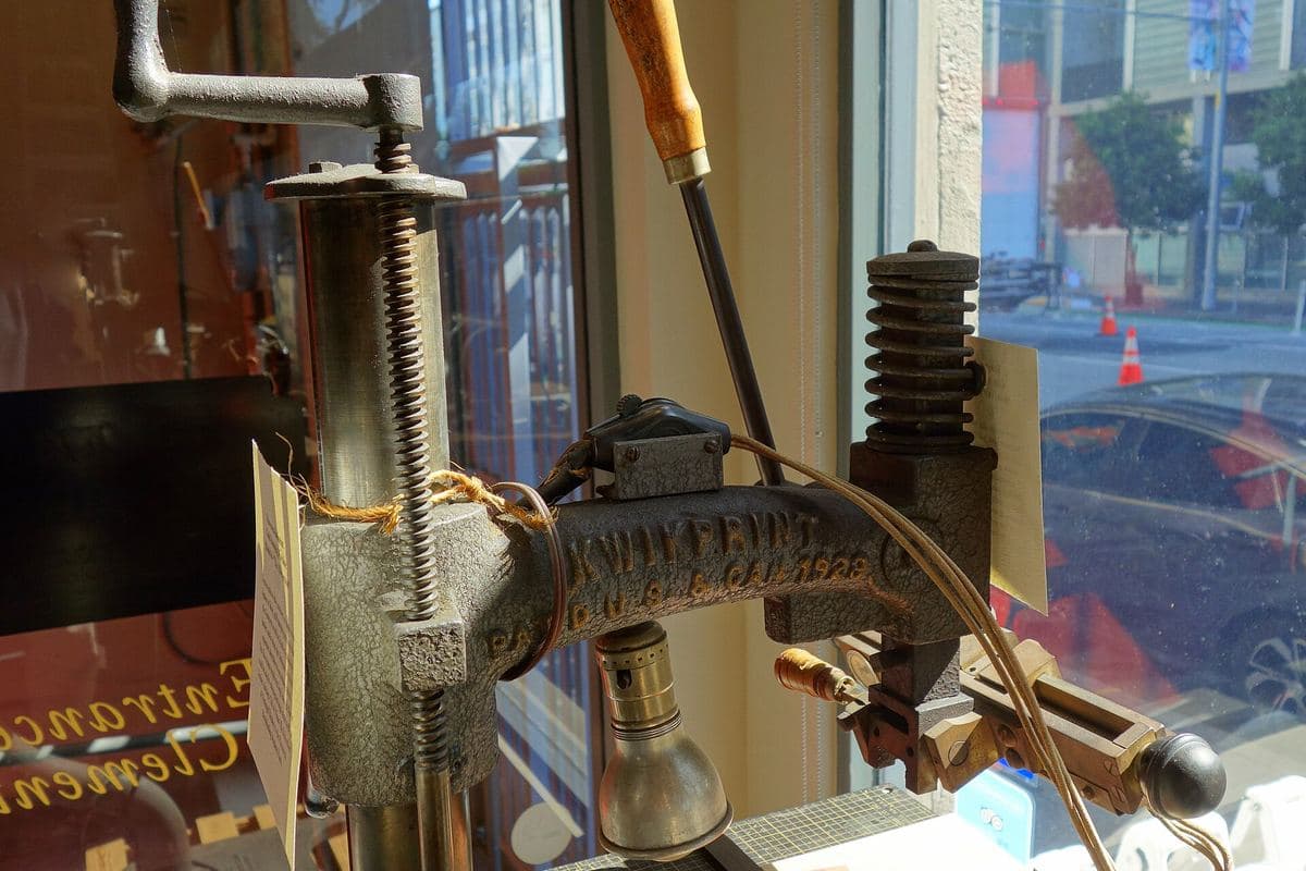 A cast-iron hand-operated gold-stamping press with a brass finishing tool and a small lamp, standing in a glazed museum window.
