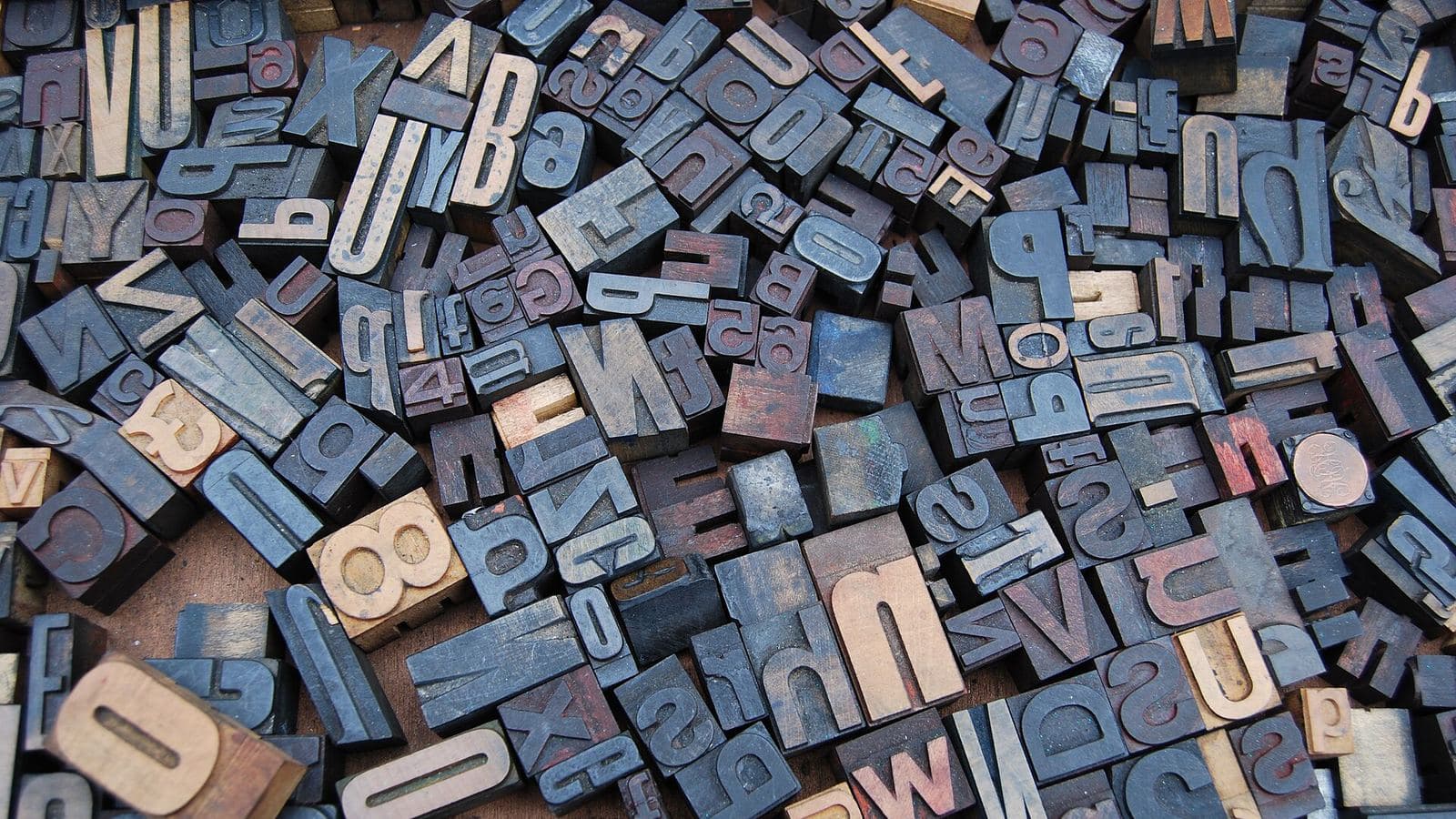 A pile of loose wooden letterpress letters and numerals on a wooden surface, some of them still carrying traces of coloured ink.